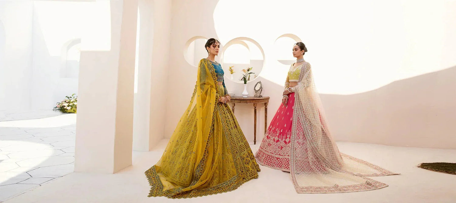 Women in elegant designer formal lehenga dresses, yellow and pink, standing in bright modern setting